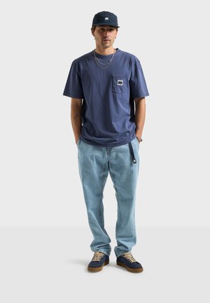 Navy cotton t-shirt with a pocket, light blue denim pants, and navy sneakers with tan soles. Accessories include a dark cap and layered necklaces.