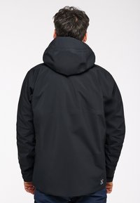 Waterdichte zwarte jas met capuchon, voorzien van een strak ontwerp, een gladde textuur en verstelbare mouwen. Subtiele branding aan de onderrand.