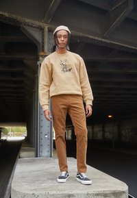 Sweatshirt bege com design gráfico, calças de veludo cotelê castanhas e ténis pretos e brancos. O sujeito posa sob um viaduto.