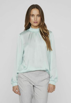 Light mint green satin blouse with long sleeves and smocked neckline. Features relaxed fit and gathered cuffs. Paired with light gray trousers.