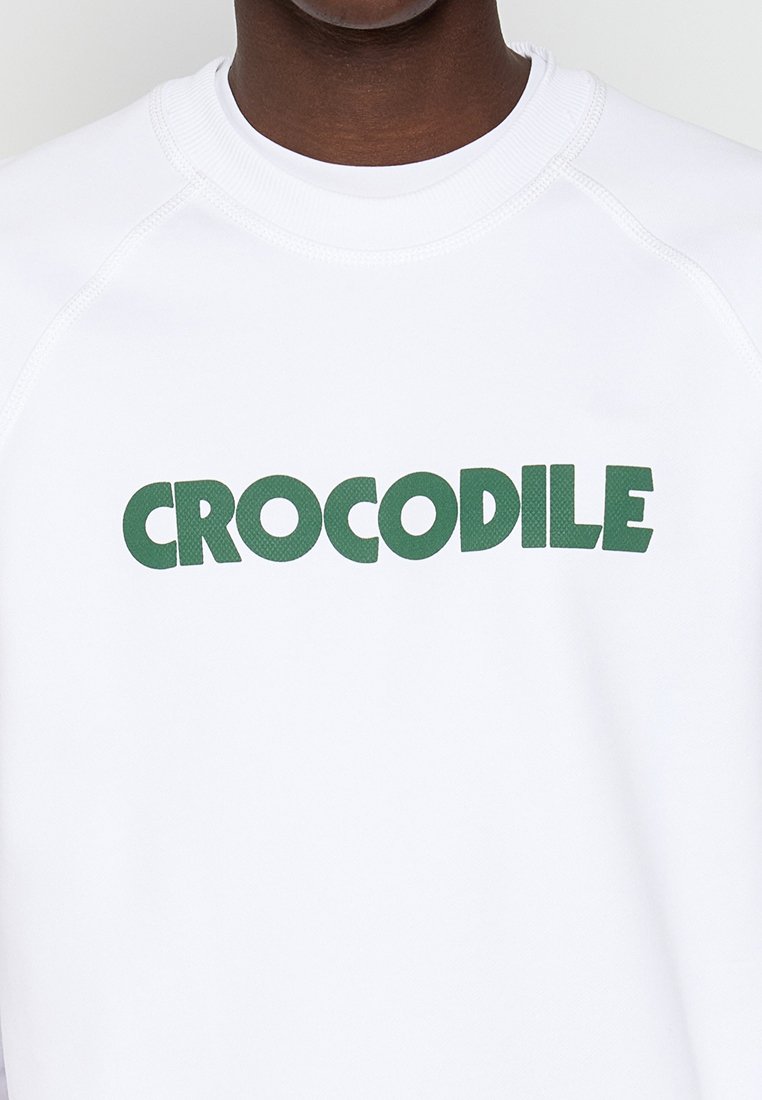 Weißes Sweatshirt mit grüner "CROCODILE"-Schrift. Verfügt über einen runden Ausschnitt und Raglanärmel, hergestellt aus einem glatten, leichten Material.