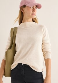 Helles beiges Strickpullover mit gerippter Textur, hochgeschlossen und mit überschnittenen Schultern. Kombiniert mit einer grünen Daunenhandtasche.