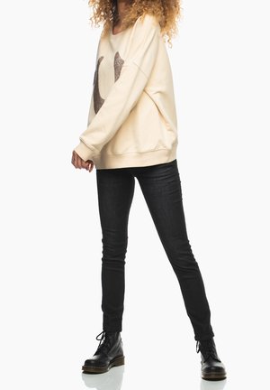 Persona con sudadera beige con un patrón oscuro, jeans ajustados negros y botas negras con cordones, de pie contra un fondo blanco.