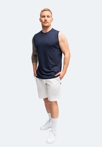Maglietta senza maniche blu navy, pantaloni corti bianchi e sneakers bianche. Presenta un tatuaggio sul braccio sinistro. Tessuto morbido con una vestibilità casual.