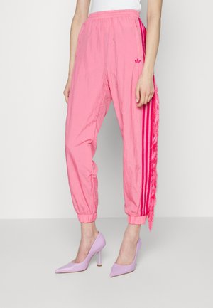 Frau trägt pinke Adidas-Trainingshose mit drei dunkelrosa Streifen und Fransen an der Seite, kombiniert mit lavendelfarbenen High Heels.