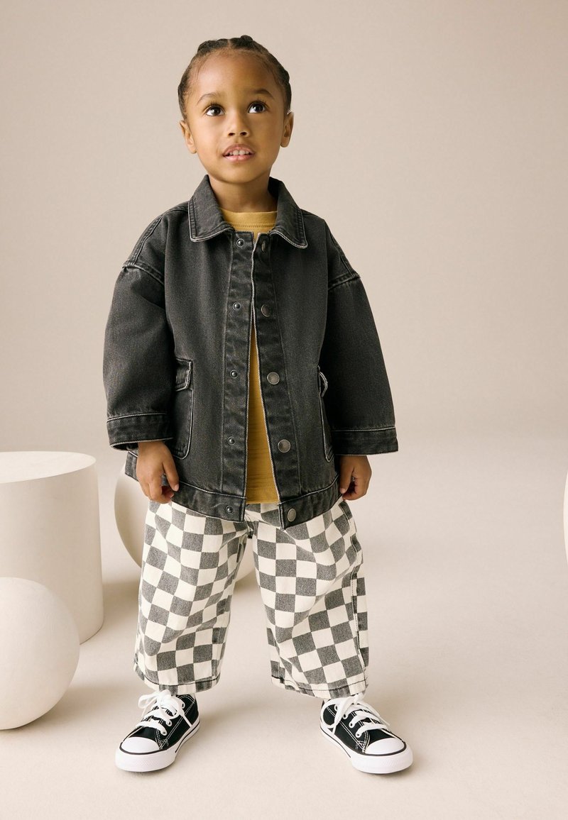 Veste en denim noire avec de grandes poches, associée à un haut jaune et un pantalon large à carreaux noir et blanc. Des baskets noires complètent l'ensemble.