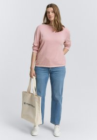Frau trägt einen pink gestreiften Pullover, blaue Jeans, weiße Sneaker und hält eine beigefarbene Tragetasche mit dem Logo "TOM TAILOR", steht vor einem weißen Hintergrund.