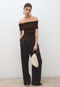 Donna che indossa un top scuro senza spalline e pantaloni larghi, tiene una borsa a tracolla in tessuto beige con manici ad anello dorati, in piedi contro un muro bianco.
