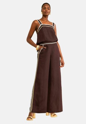 Kvinde iført mørkebrune bukser med vide ben og matchende ærmeløs top med hvid bølget kant, accessoriseret med guldarmbånd og hælede sandaler.