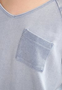 T-shirt azzurro chiaro con scollatura a V e una tasca in denim testurizzato. Il tessuto appare morbido e leggermente elasticizzato, con dettagli di cuciture sottili.