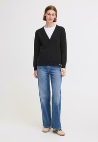 Cardigan noir en laine avec un décolleté en V, boutons sur le devant et détail de boutons sur les côtés, associé à un jean à jambes larges en denim délavé et des chaussures beiges.