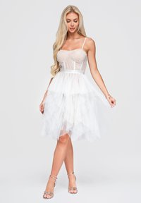 Femme blonde en robe corset en dentelle blanche avec une jupe en tulle à étages et des sandales métalliques à talons hauts, debout devant un fond uni.