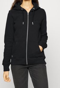 Sudadera negra con cremallera hecha de tejido suave, con capucha forrada, cordones, dos bolsillos frontales y puños y dobladillo acanalados.