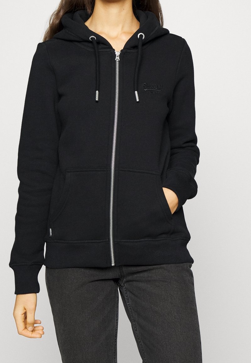 Sudadera negra con cremallera hecha de tejido suave, con capucha forrada, cordones, dos bolsillos frontales y puños y dobladillo acanalados.