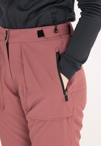 Vaaleanpunaiset softshell-housut, joissa on mustat vetoketjut ja taskut. Housuissa on suora leikkaus ja teksturoitu, joustava kangas, joka tarjoaa mukavuutta ja liikkuvuutta.