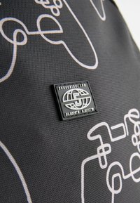 Schwarze Stofftasche mit weißen abstrakten Linienmustern. Beinhaltet ein schwarzes Etikett mit eingeprägtem Text und Koordinaten auf einem rechteckigen Patch.