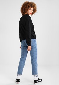 Fekete hosszúujjú póló, világoskék farmernadrág és fekete sportcipő. Göndör haj, semleges háttérben állva. Csíkos fehér zoknik.