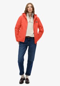 Chaqueta impermeable roja con capucha, con cremallera en la parte delantera, combinada con un forro polar gris claro y jeans de mezclilla azul, llevada con mocasines marrones y calcetines blancos.