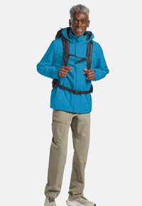 Giacca impermeabile blu con cappuccio, dotata di cinturini regolabili e una fibbia. Abbinata a pantaloni beige chiaro e scarpe da trekking grigie.