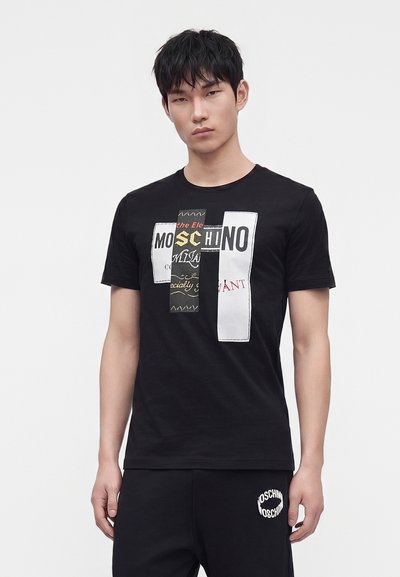 Άνδρας που φοράει μαύρο μπλουζάκι Moschino με αφηρημένο λογότυπο και μαύρο παντελόνι με το λογότυπο Moschino στο αριστερό μηρό, στέκεται μπροστά σε απλό φόντο.
