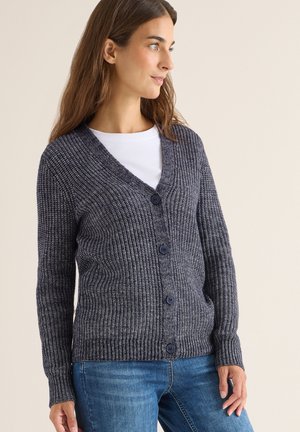 Tummansininen neulecardigan, jossa on V-pääntie, napit edessä, ribbit kädenteitse ja teksturoitu kangas. Yhdistetty valkoisen paidan ja farkkujen kanssa.