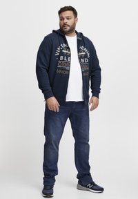 Felpa con zip blu navy con stampa, abbinata a una maglietta bianca e jeans blu scuro. Presenta tasche laterali e polsini a coste.