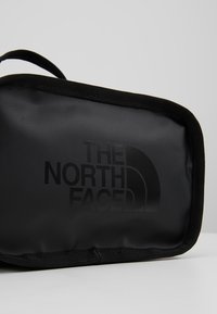 Sac rectangulaire noir avec une poignée et le logo The North Face imprimé en noir foncé sur le devant sur fond gris clair.