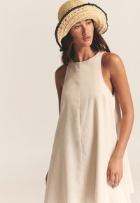 Hellbeiges ärmelloses Kleid mit lockerem Schnitt, einem hohen Ausschnitt und seitlichen Aussparungen. Getragen mit einem gewebten Strohhut mit schwarzen Akzenten.