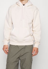 Sudadera de color beige claro con un bolsillo frontal y capucha con cordón, hecha de tela suave. Presenta puños y dobladillo acanalados. Combinada con pantalones cargo de color oliva.