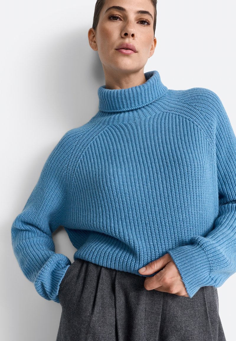 Pulover albastru tricotat cu guler înalt, textură cu nervuri și croială lejeră. Asortat cu pantaloni gri plisati, cu mâinile odihnindu-se pe talie.