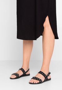 Sandalias planas negras de cuero con tachuelas, que cuentan con dos correas en el pie y una correa ajustable en el tobillo. Suela lisa, diseño minimalista.