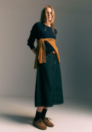 Un pull bleu marine superposé avec un pull moutarde, associé à une longue jupe en denim. Des chaussures brunes avec des lacets, des chaussettes foncées et une taille ceinturée.