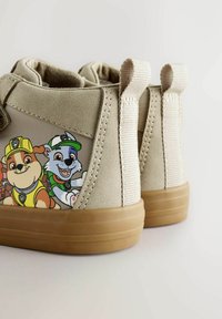 Beige Kleinkinderschuhe mit braunen Sohlen zeigen auf der Seite animierte Hundfiguren in Bau- und Recycling-Outfits.