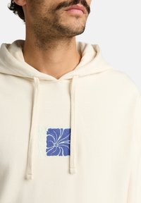 Haut à capuche crème avec une poche avant, doté d'une broderie florale bleue sur un patch carré et d'un cordon de serrage. Texture douce.
