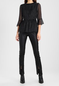 Donna che indossa una blusa nera trasparente a pois con maniche a volant e pantaloni di pelle neri con fessure anteriori alla caviglia e stivali neri.