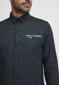 Temno modra srajca na gumb z teksturirano tkanino, ki ima žep na prsih z svetlo modrim robom in majhnim rdečim, belim in modrim logotipom.
