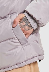 Svetlofialová péřová bunda s elastickými manžetami. Ruka v kapse ukazuje hladký povrch a detaily šití. Viditeľné tatto na zápästí.