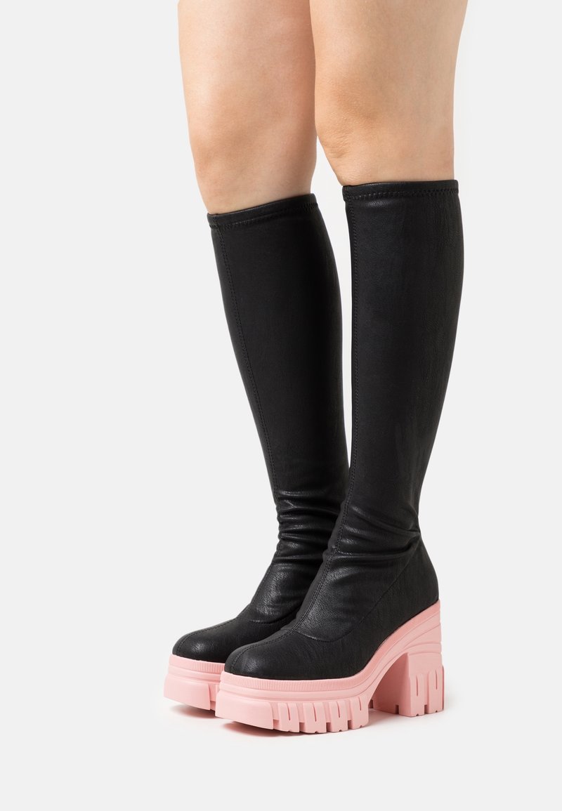 Bottes noires en similicuir montant jusqu'au genou avec un design décontracté, une épaisse semelle plateforme rose et une texture rainurée qui crée un contraste et met en valeur la chaussure.