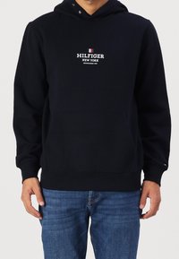 Sudadera de algodón azul marino con bolsillo frontal, capucha con cordones y texto del logo en blanco. Puños y dobladillo acanalados. Combinada con jeans de mezclilla azules.