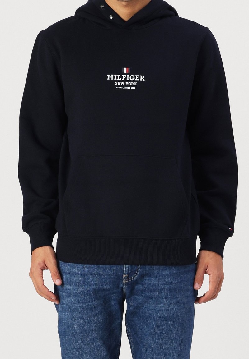 Sudadera de algodón azul marino con bolsillo frontal, capucha con cordones y texto del logo en blanco. Puños y dobladillo acanalados. Combinada con jeans de mezclilla azules.