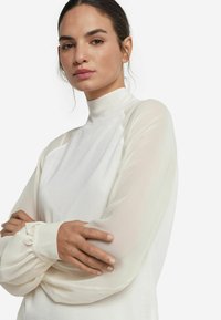 Top bianco a maniche lunghe con collo alto, caratterizzato da un corpo liscio e uniforme e maniche trasparenti e svolazzanti con polsini arricciati.