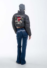 Schwarze Pufferjacke mit einem kurzen Design, die ein buntes Totenkopf-Motiv zeigt. Eine Hose aus blauen Jeans mit bestickten Details und Logo.