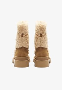 Botines de felpa beige forrados de piel, con una suela de goma marrón resistente, que presentan un panel trasero de ante suave y detalles de correa ajustable.