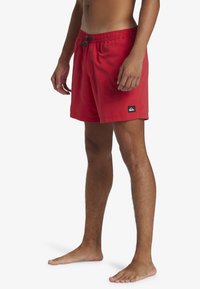 Piros fürdőshorts, könnyű szövetből készült, elasztikus derékpánttal és húzózsinórral, az alján egy kis fekete logópáccsal.