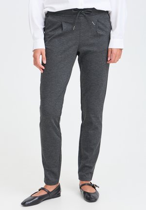 Person trägt dunkelgraue Hose mit Kordelzug, weißes Langarmshirt und schwarze flache Schuhe und steht vor einem schlichten Hintergrund.