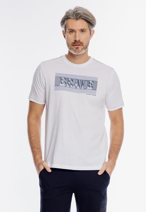 Mężczyzna z siwymi włosami ubrany w białą koszulkę z napisem „BRAVE” i ciemne spodnie, stoi z rękami w kieszeniach na białym tle.