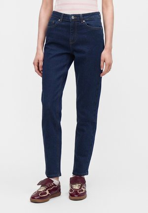Person iført mørkeblå jeans med lige ben, hvide sokker og mørkerøde sneakers med beige snørebånd, stående mod en ensfarvet baggrund.