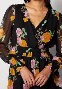 Femme portant une robe noire à imprimé floral coloré, col cache-cœur, manches longues transparentes, taille et poignets smockés, mains jointes sous la taille.