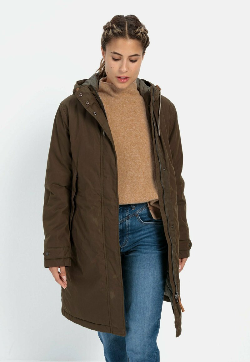 Olivgrüner Parka mit Kapuze, ausgestattet mit einem Reißverschluss und Druckknopfverschluss, verstellbaren Kordeln und sichtbaren Seitentaschen. Getragen über einem hellbraunen Pullover.