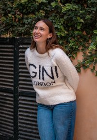 Smiling woman in white fuzzy sweater with "GINI LONDON" text, blue jeans, standing against wall with green leafy vines.